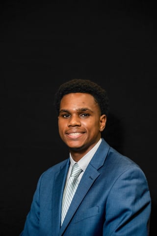 Professional headshot of a smiling man in a blue blazer and striped tie against a black background