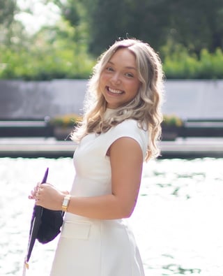 Young woman with blonde wavy hair smiling at camera wearing white dress and holding purple oar by water