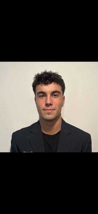 Portrait of a young man with dark curly hair wearing a black blazer and shirt against a neutral beige background
