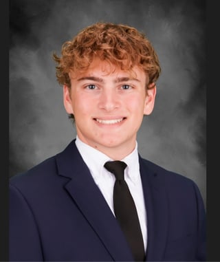 Young man with curly reddish-brown hair smiling in professional headshot wearing navy blazer, white shirt, and black tie
