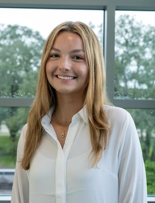 Young woman with long blonde hair wearing a white button-up shirt, smiling at the camera in front of a window with trees visible outside